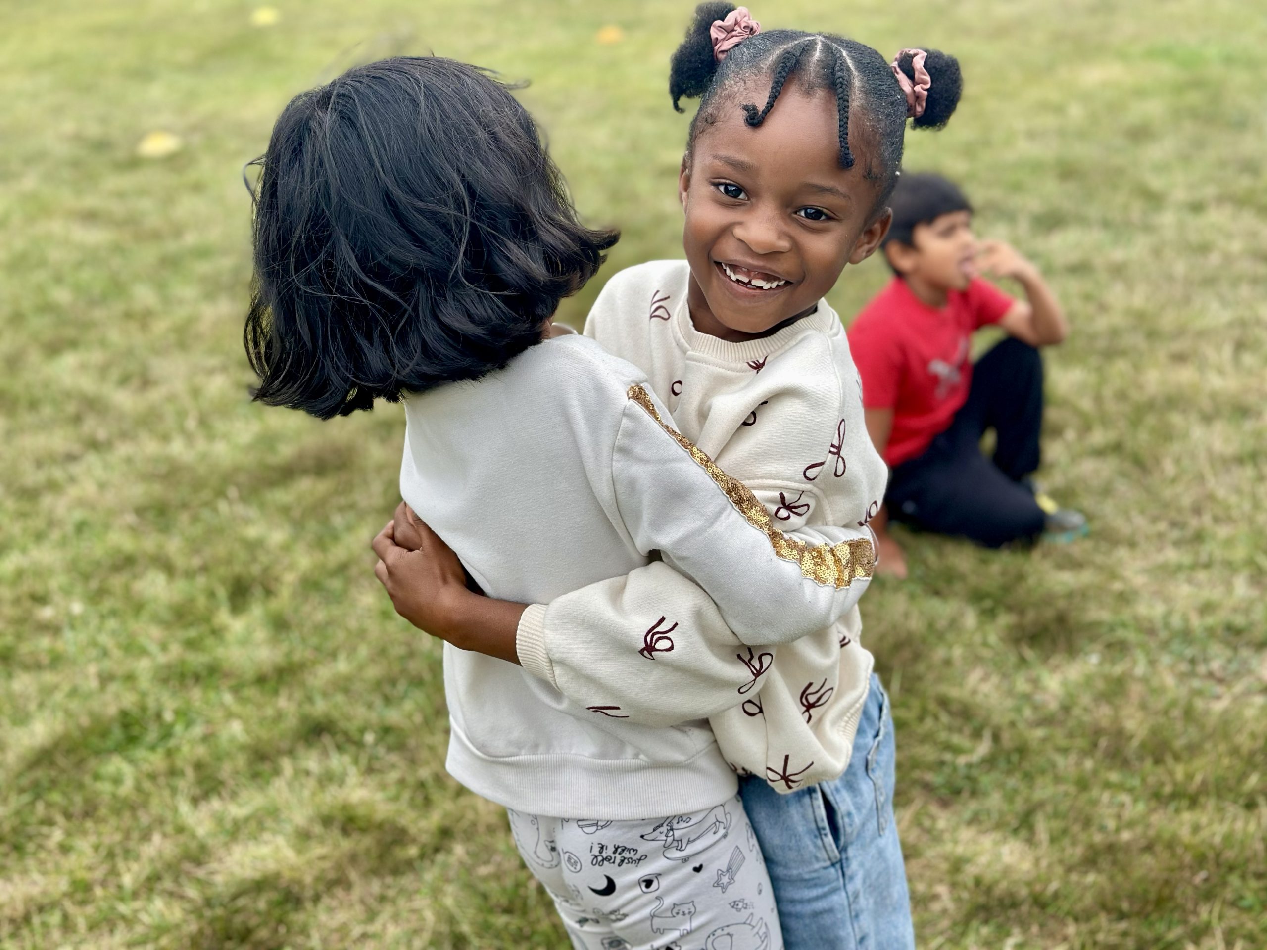 Two young children hug and smile outdoors during the Town of Ajax's Playgrounds Program while another child sits on the grass nearby.