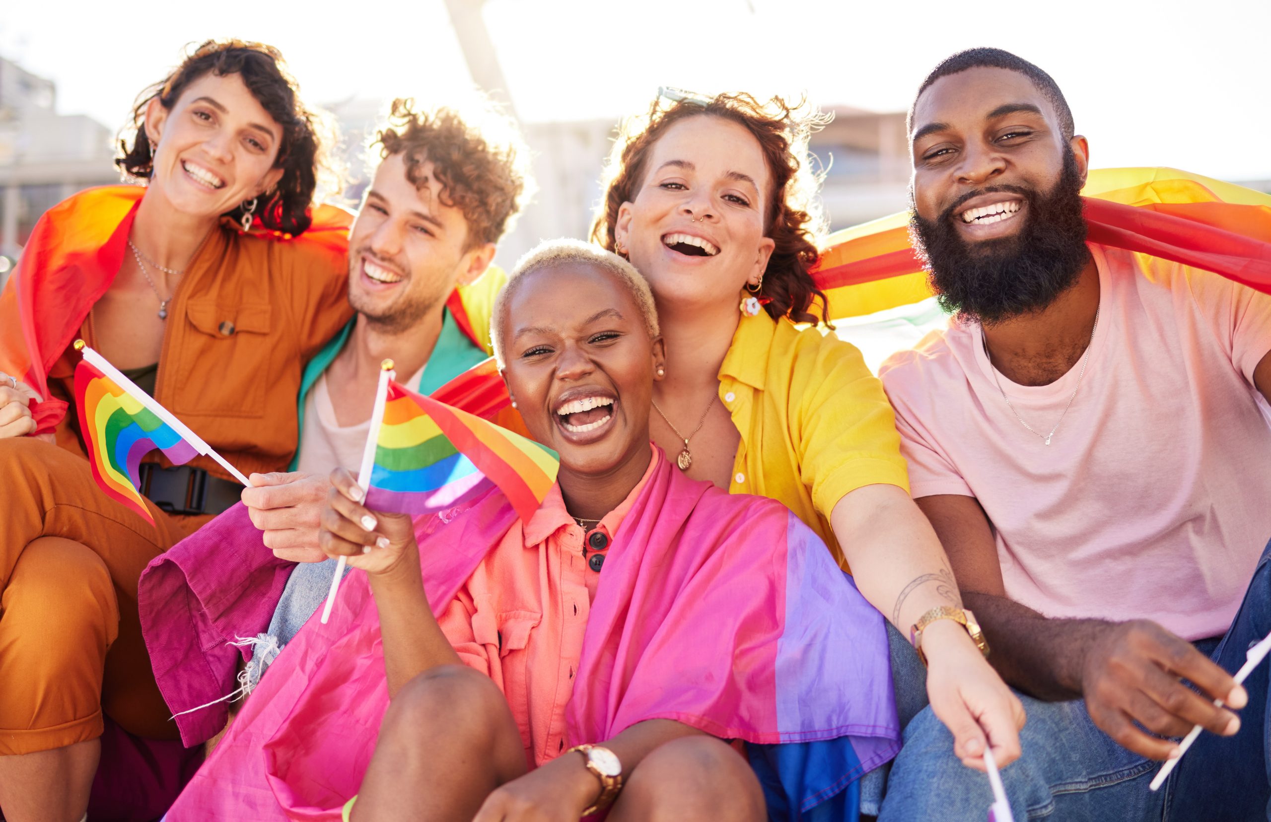 Five people sitting outdoors, smiling and holding rainbow pride flags, with rainbow flags draped around their shoulders.