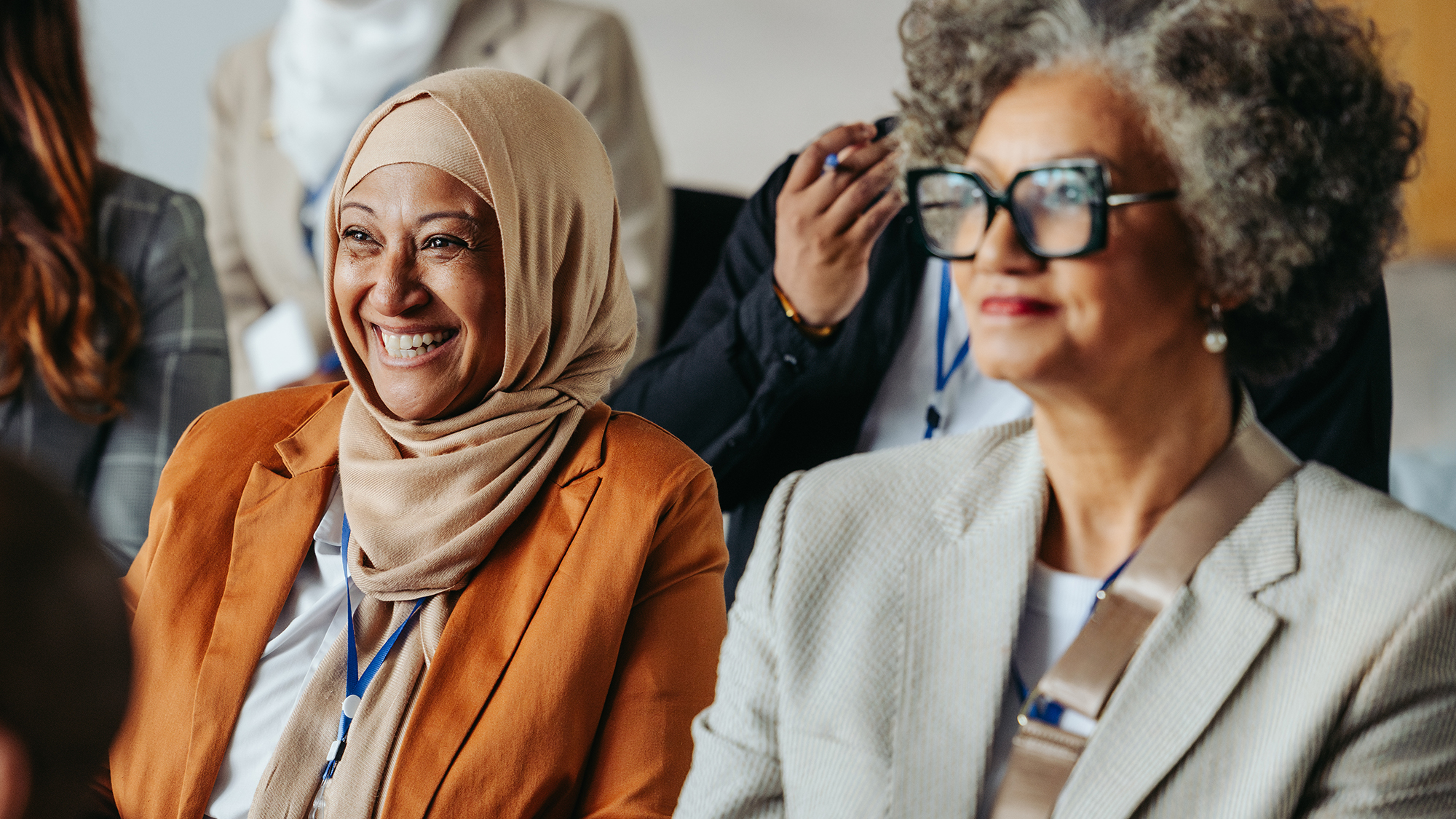 A diverse group of professionals, including a smiling Muslim woman, listen to a presentation.