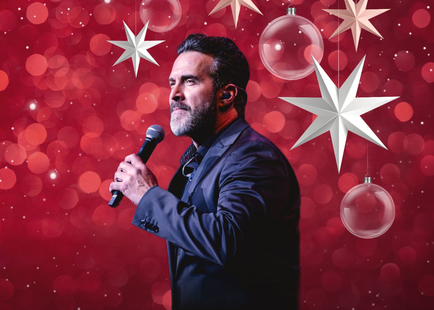 A singer dressed in a suite holding a microphone with a red backdrop behind him with stars and Christmas Balls