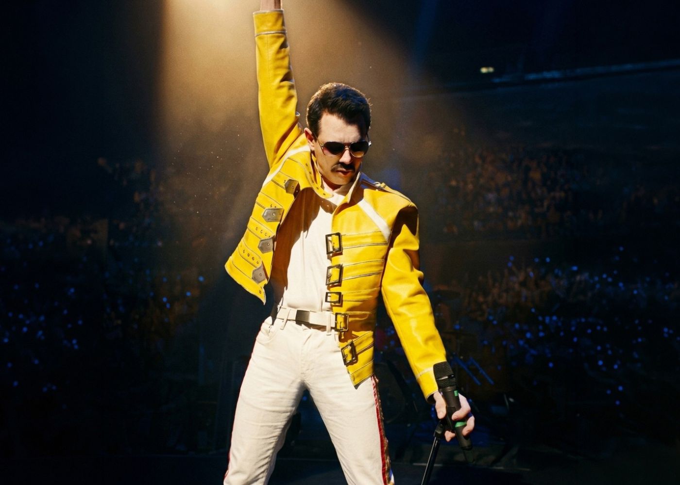 A singer on stage in a yellow jacket and white pants, holds a microphone in his left hand on stage with his right arm reaching in the air