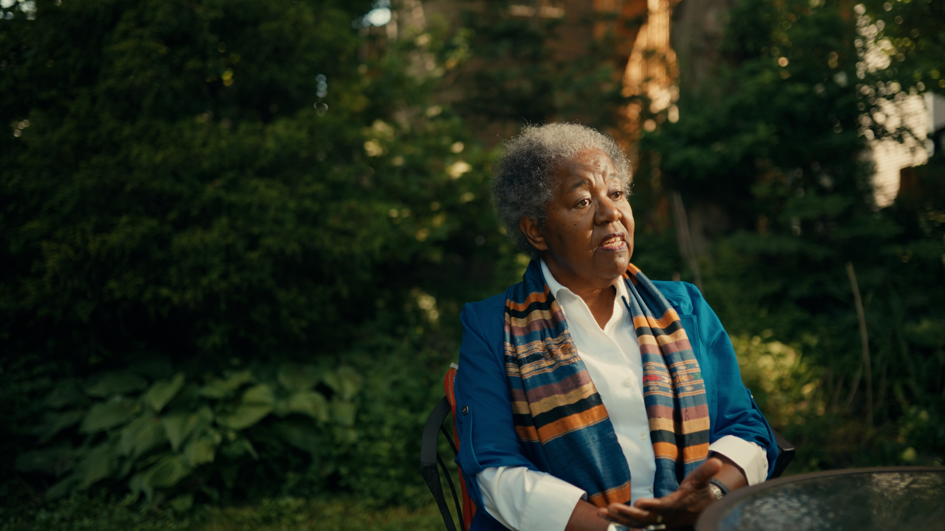 A women sitting outside on a fall day weaing a blue jacket, white button up top and a stripped scarf with trees in the background