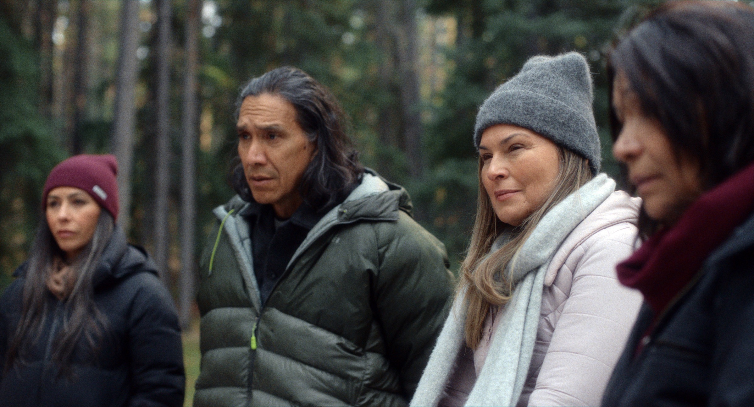 4 people standing outside in a wooded area dressed in winter jackets and 2 with hats.