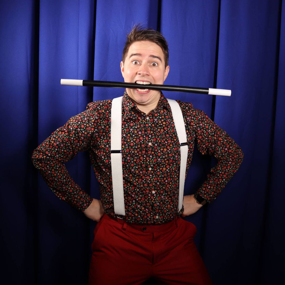 A magician wearing suspenders poses playfully with a magic wand held between his teeth, against a blue stage curtain