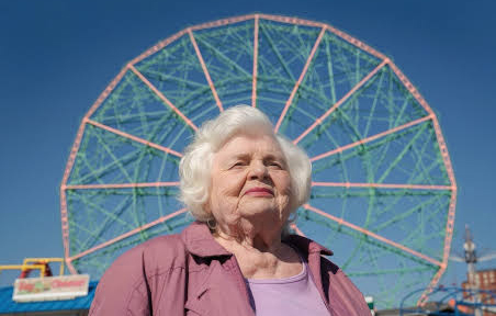 An elderly lady wearing a pink jacket stands in front of a ferris wheel.