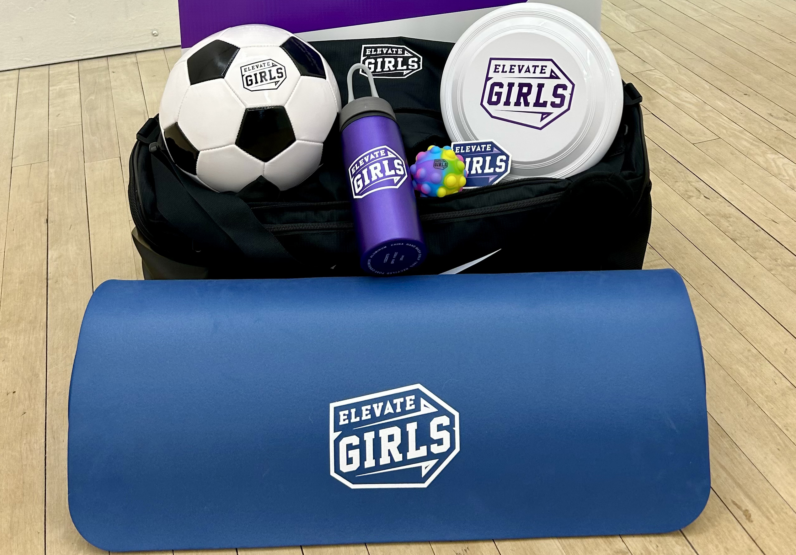 A prize package consisting of a Nike duffel bag, soccer ball, water bottle, yoga mat, sticker, frisbee and pop it ball are arranged on a gym floor in front of an informational Elevate Girls poster.