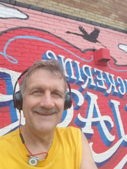 A man with headphones stands in front of the Pickering Village mural.